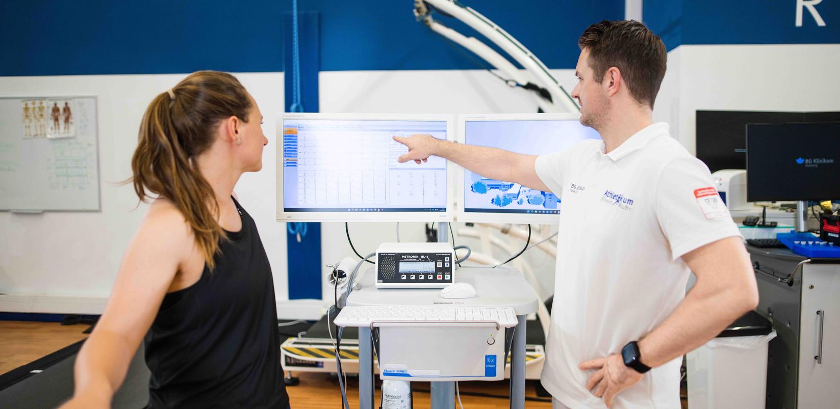 Der Arzt erklärt der Patientin ihre Gesundheitswerte anhand zwei Monitoren