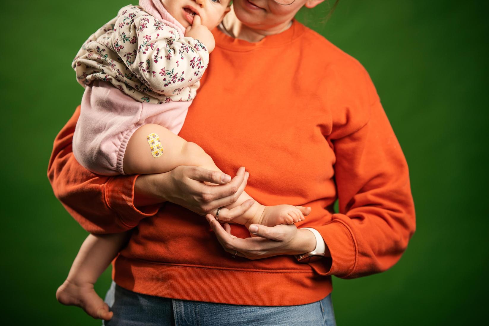 Eine Mutter im orangefarbenen Pulli hält ihr Baby auf dem Arm. Das Mädchen hat ein Pflaster auf dem Oberschenkel.