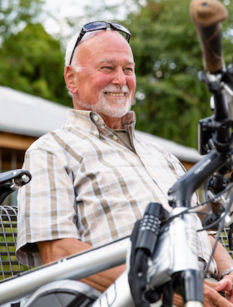 Älterer Herr sitzt lächelnd auf einer Bank mit einem Fahrrad vor sich stehend.