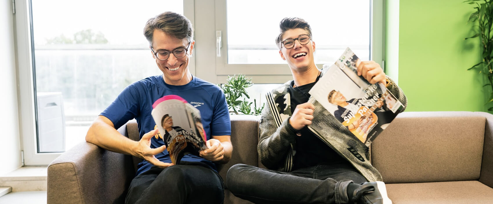 Zwei Männer sitzen lachend auf einem Sofa und blättern im Bergische Krankenkasse Kundenmagazin